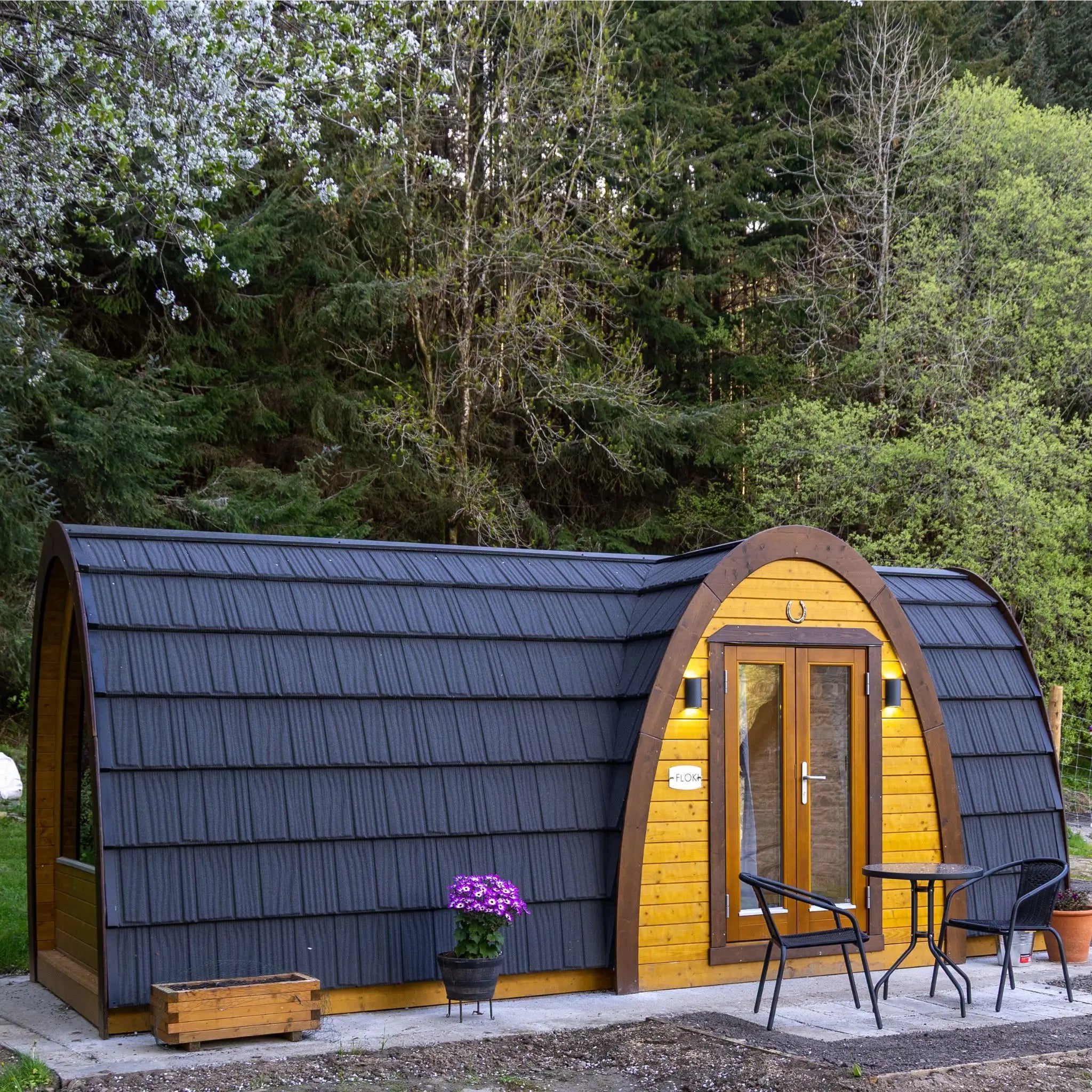 A small arched cabin or pod structure with dark charcoal metal siding and warm yellow wood paneling, shown outdoors on a patio with a small table, chairs, and a potted plant. Trees and shrubs fill the background.