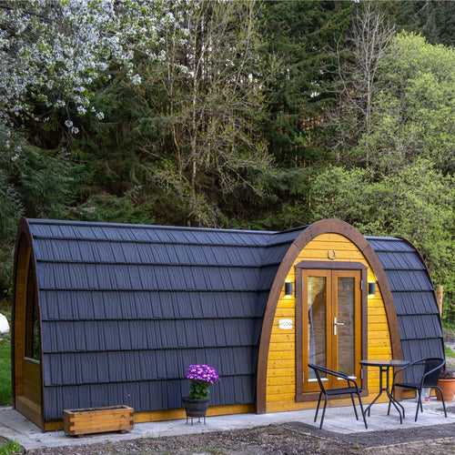 A small arched cabin or pod structure with dark charcoal metal siding and warm yellow wood paneling, shown outdoors on a patio with a small table, chairs, and a potted plant. Trees and shrubs fill the background.