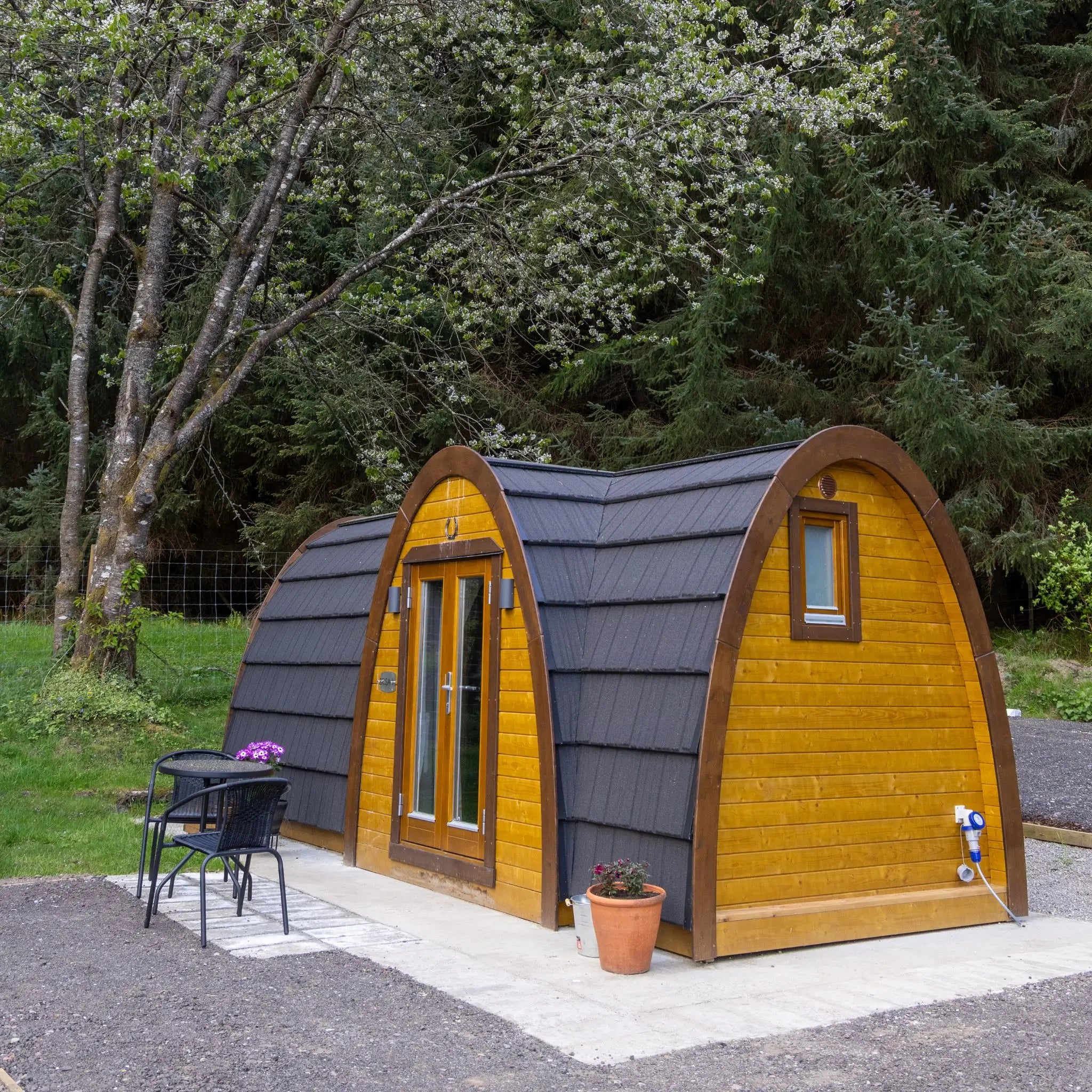 A small arched wooden cabin or playhouse with yellow wood siding, dark gray shingled roof, and a double door, shown outdoors on a patio with trees and grass in the background.