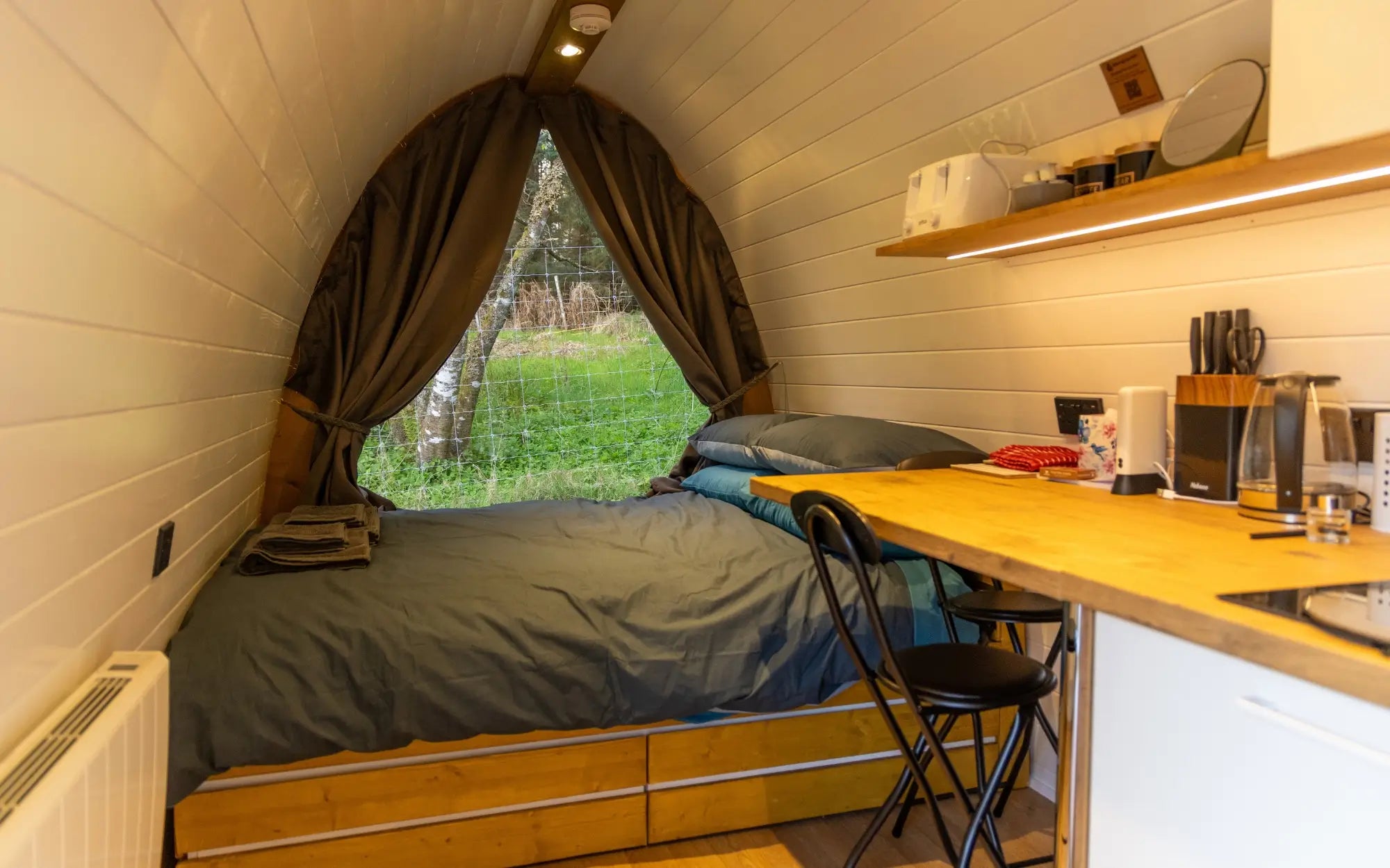 A cozy compact camper bed with a built-in kitchenette, featuring a wooden bed frame, gray bedding, a wooden desk, and a window with dark curtains looking out onto green grass.