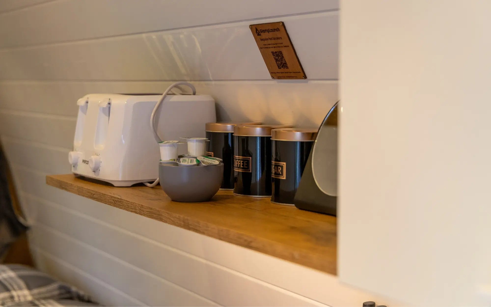 A wooden shelf mounted on a white shiplap wall, holding a white toaster, a small gray bowl, and several dark canisters with gold lids. A small wooden sign is attached above the shelf.