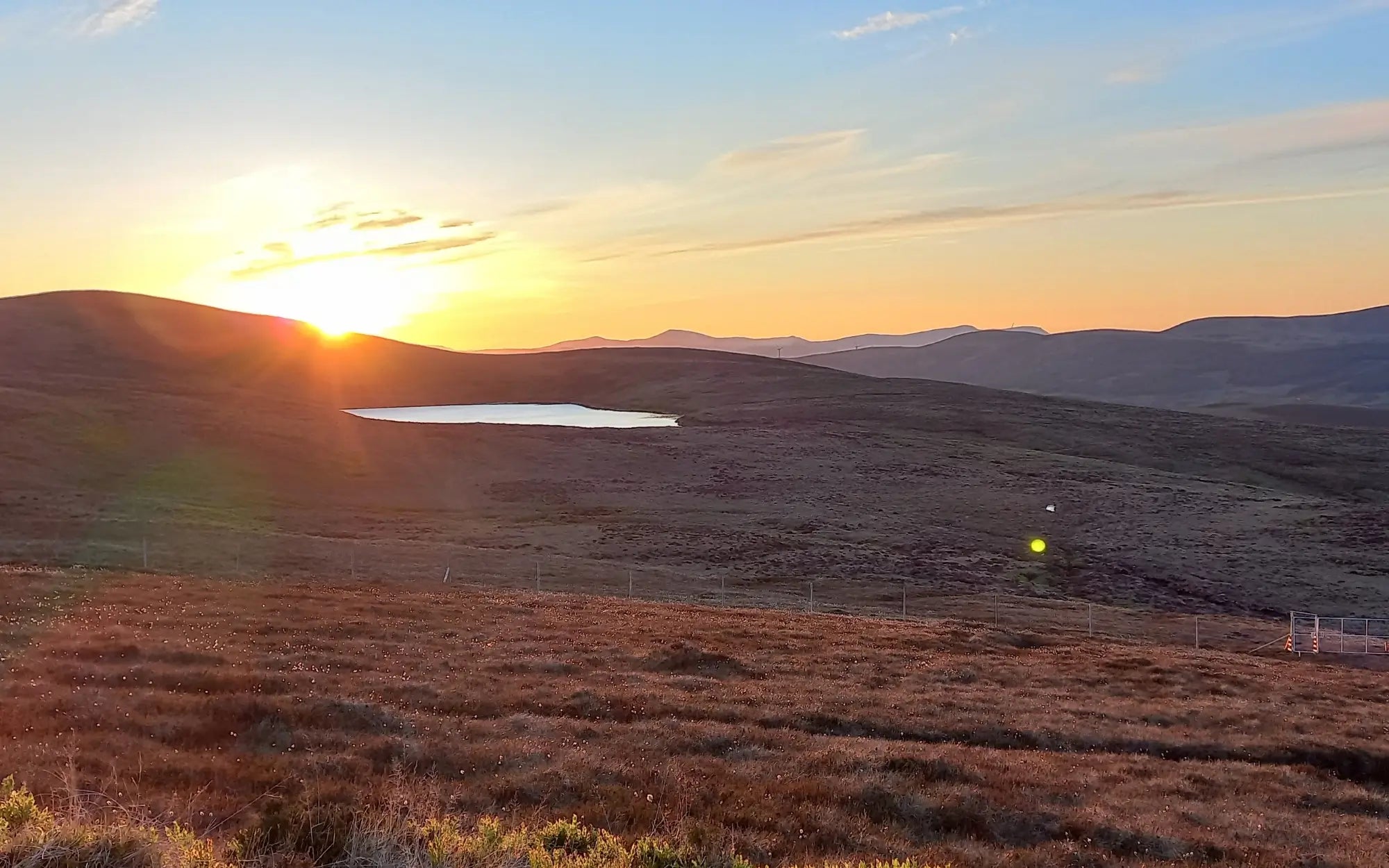 A scenic landscape photograph of a sunset over rolling hills and a small lake, with golden light on the horizon and a blue sky above.